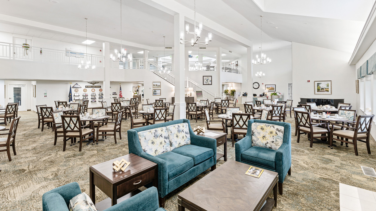 A spacious, bright dining area with many tables and chairs, teal upholstered seating in the foreground, chandeliers, and large windows letting in natural light. The room has light walls and an open, airy layout.