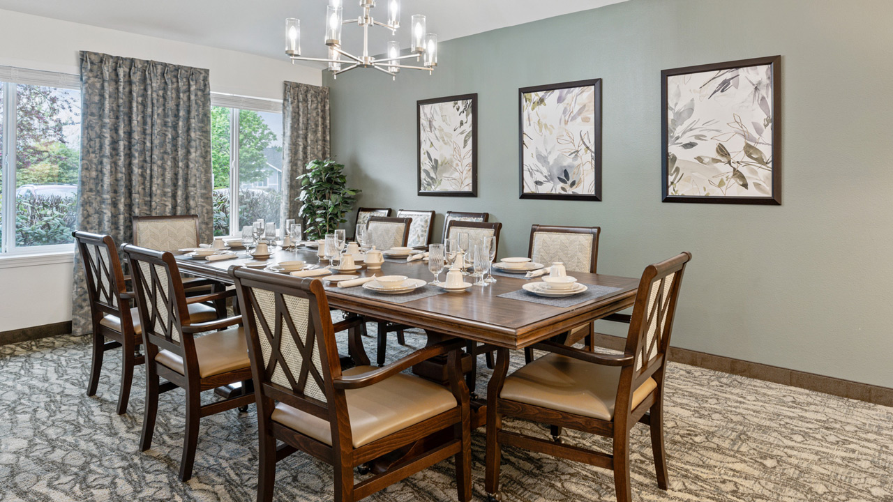 Elegant dining room with a large wooden table set for ten, cushioned chairs, neutral décor, three botanical art pieces on a light green wall, patterned carpet, large windows with drapes, and a modern chandelier above.