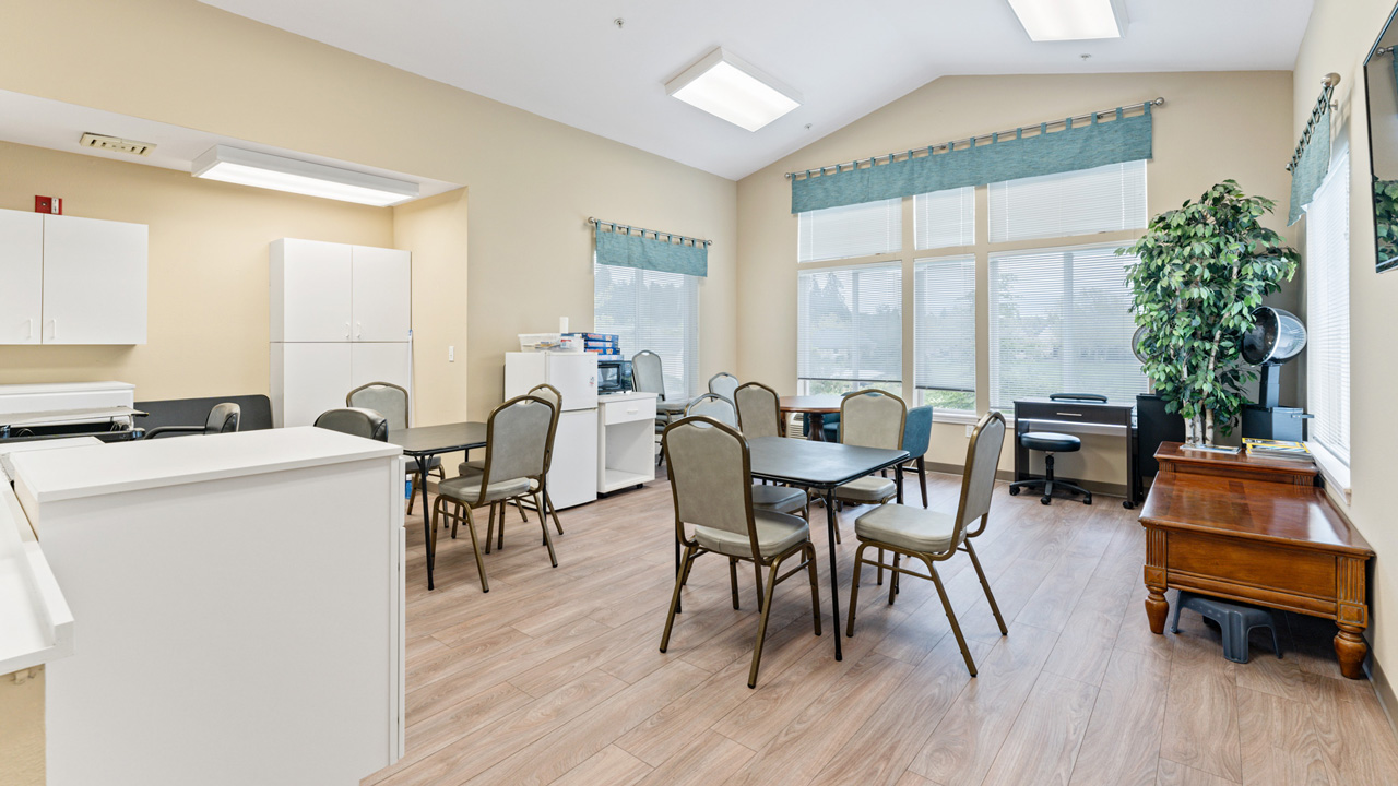 A bright, clean room with several tables and chairs, large windows with green valances, white cabinets, a printer, a file organizer, and a tall potted plant on wooden flooring.