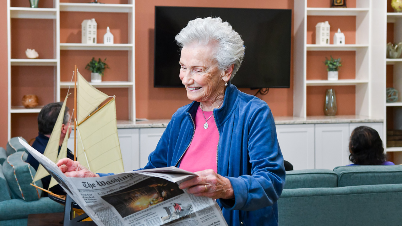 An elderly woman with short gray hair, wearing a blue jacket and pink shirt, smiles while reading The Washington Post newspaper in a cozy, well-lit living room with shelves and a model sailboat in the background.