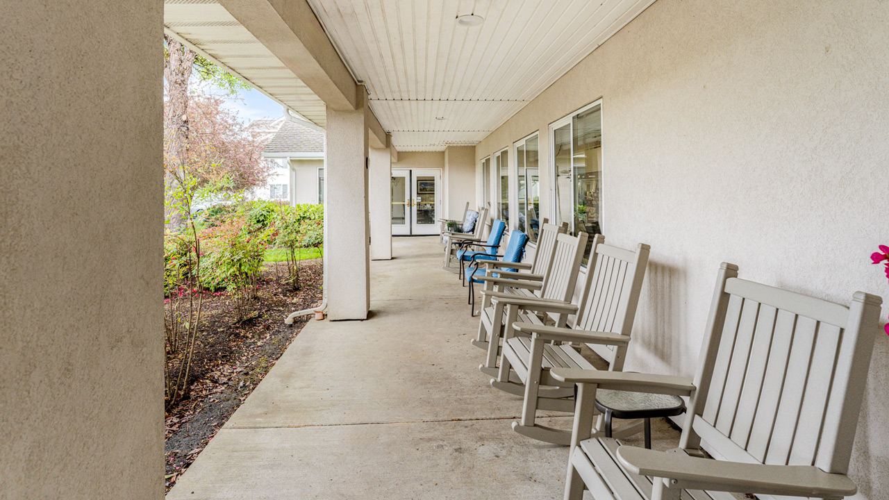 A covered outdoor walkway with several white and blue rocking chairs lined up along a beige building with large windows, overlooking a landscaped garden area.
