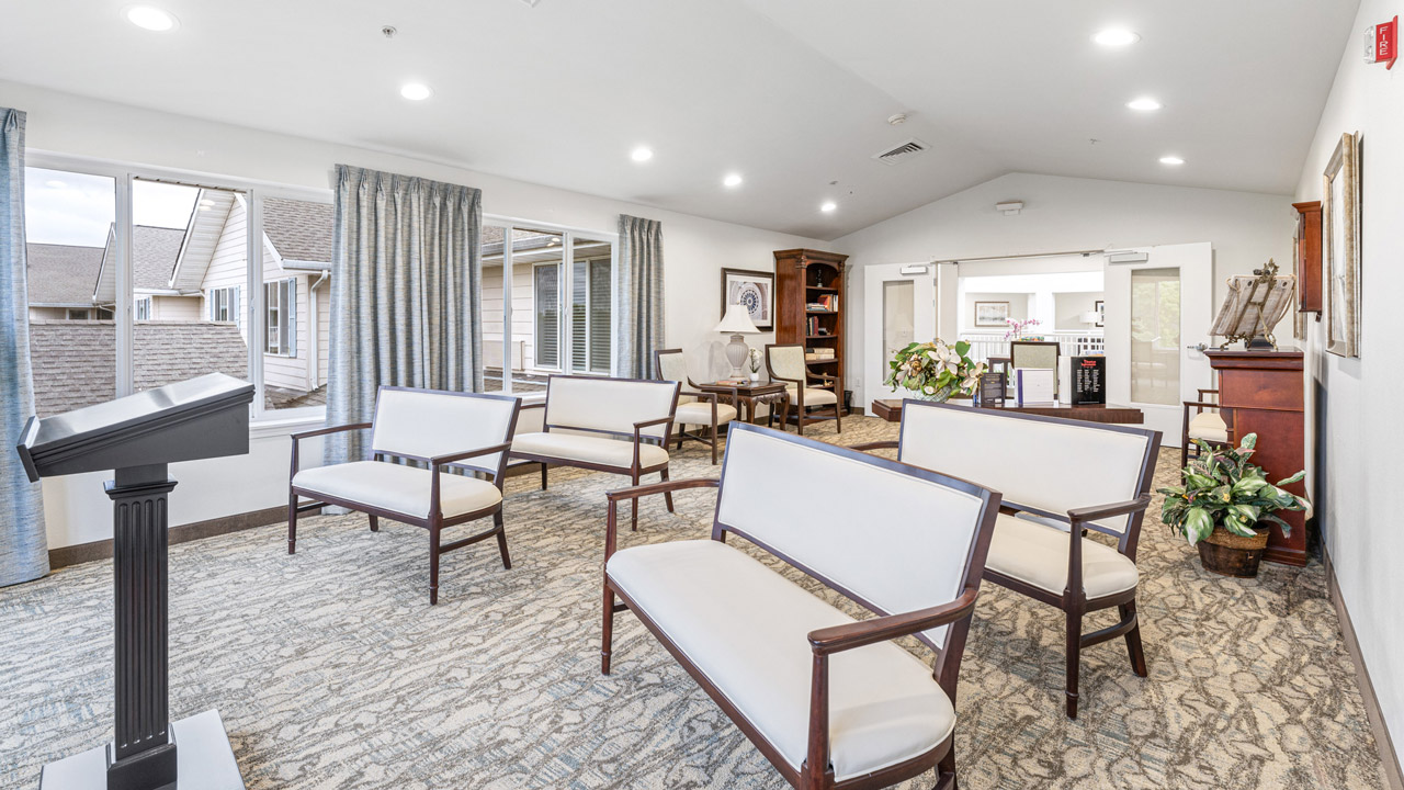 A bright, spacious room with white cushioned benches, a podium, bookshelves, large windows with curtains, plants, and neutral decor, set up for a small gathering or meeting.