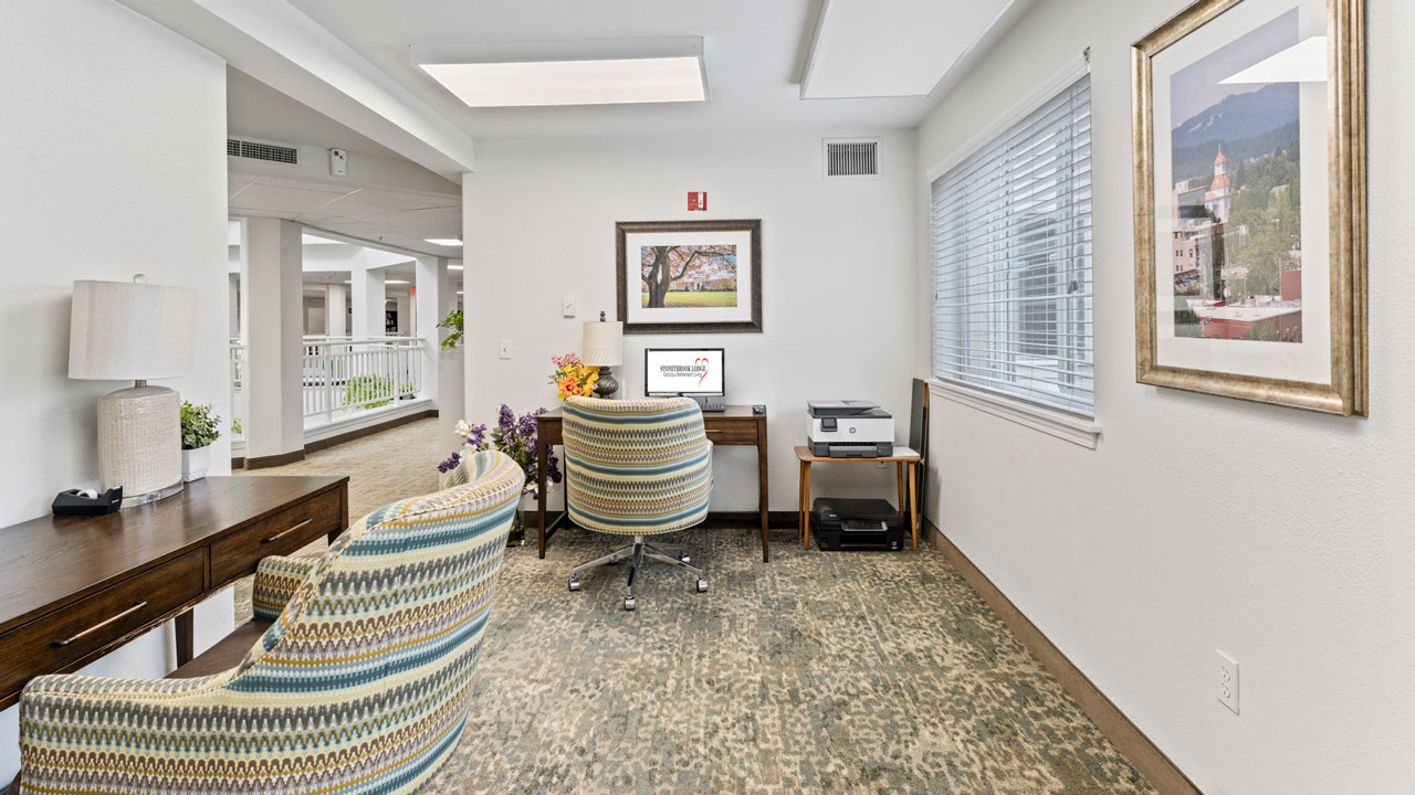 A small office area with two patterned chairs, a wooden desk, a computer, a printer, a lamp, and framed artwork on the walls. Large windows let in natural light, and the space looks tidy and inviting.
