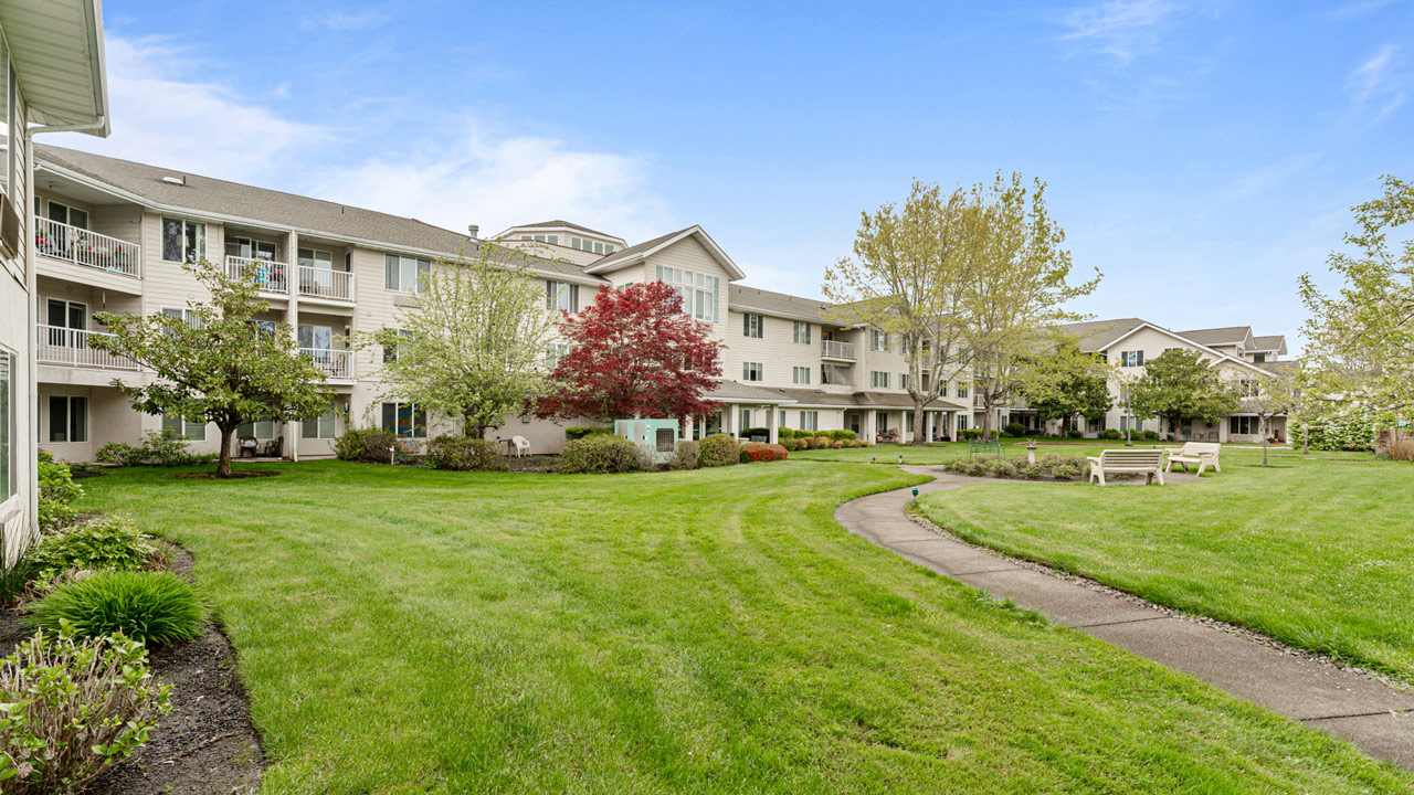A well-maintained apartment building with balconies overlooks a green lawn, trees, shrubs, and a curved pathway with benches under a partly cloudy blue sky.