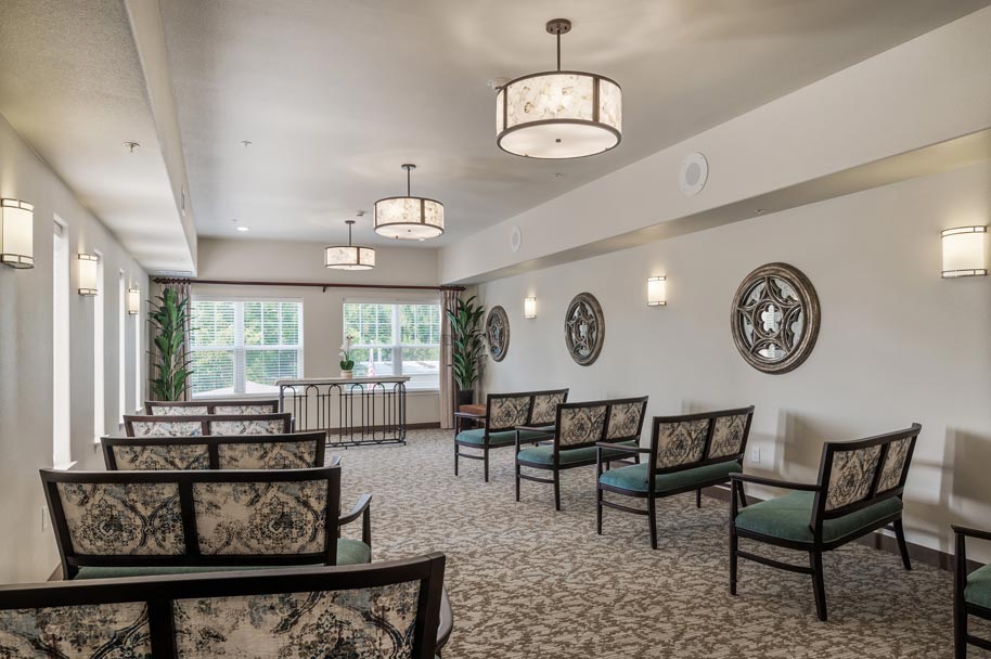 A well-lit room with patterned chairs arranged in rows facing a window. The walls are decorated with circular art pieces and wall sconces. A table with plants is in front of the window, and the ceiling has several large, round light fixtures.