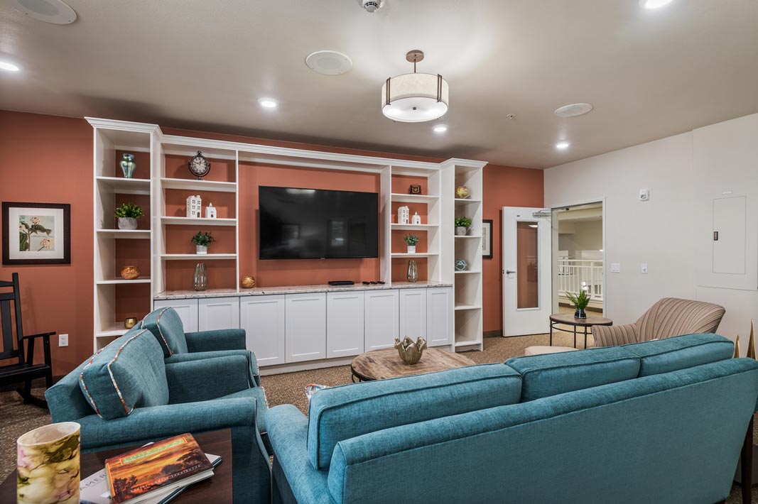 A cozy living room with teal sofas, a beige armchair, a dark wooden coffee table, and a large flat-screen TV mounted on a white built-in shelving unit. Shelves display decorative items, and the walls are painted a warm reddish-brown. Overhead lighting is on.