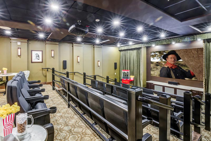 A small, cozy movie theater with a few rows of comfy seats, popcorn on tables, a popcorn machine, and a large screen displaying a movie scene. The walls are adorned with framed photos, and the ceiling has modern light fixtures.