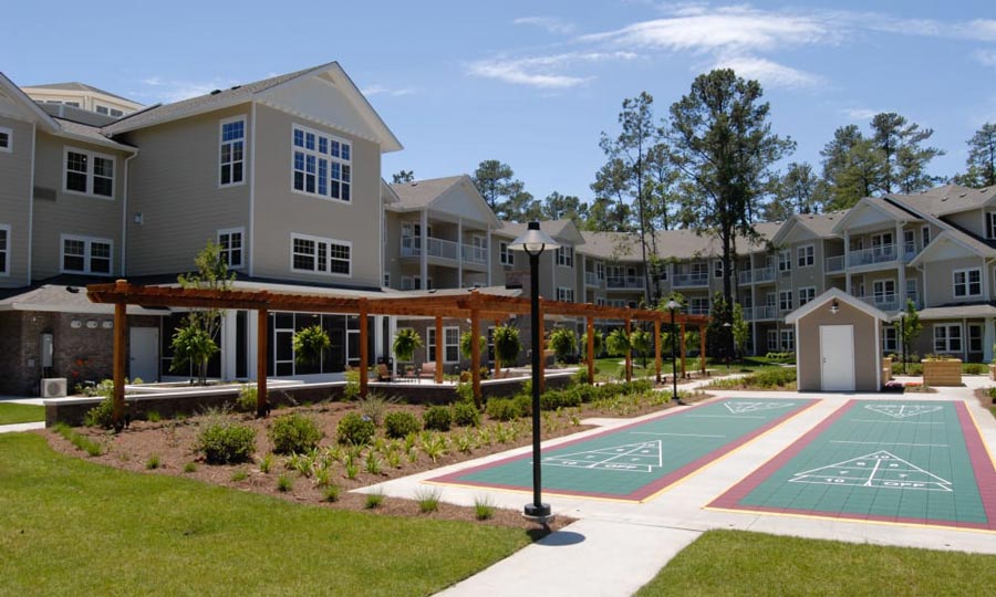 An outdoor view of a residential complex featuring modern, multi-story buildings with balconies. The courtyard includes pathways, landscaped greenery, and two shuffleboard courts. There is also a wooden pergola and a small white utility shed.