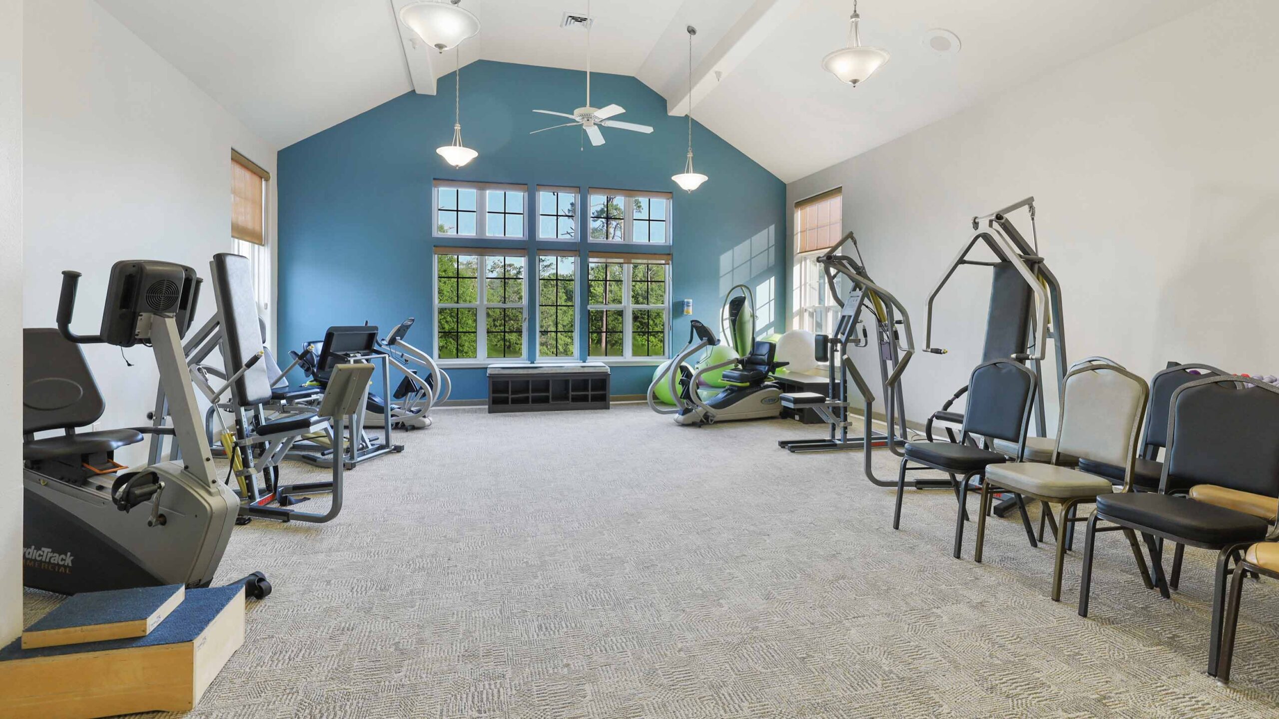A spacious, well-lit gym with exercise machines along the walls, large windows, light beige carpet, and blue accent wall at the far end. Stacked chairs are placed on the right side of the room.