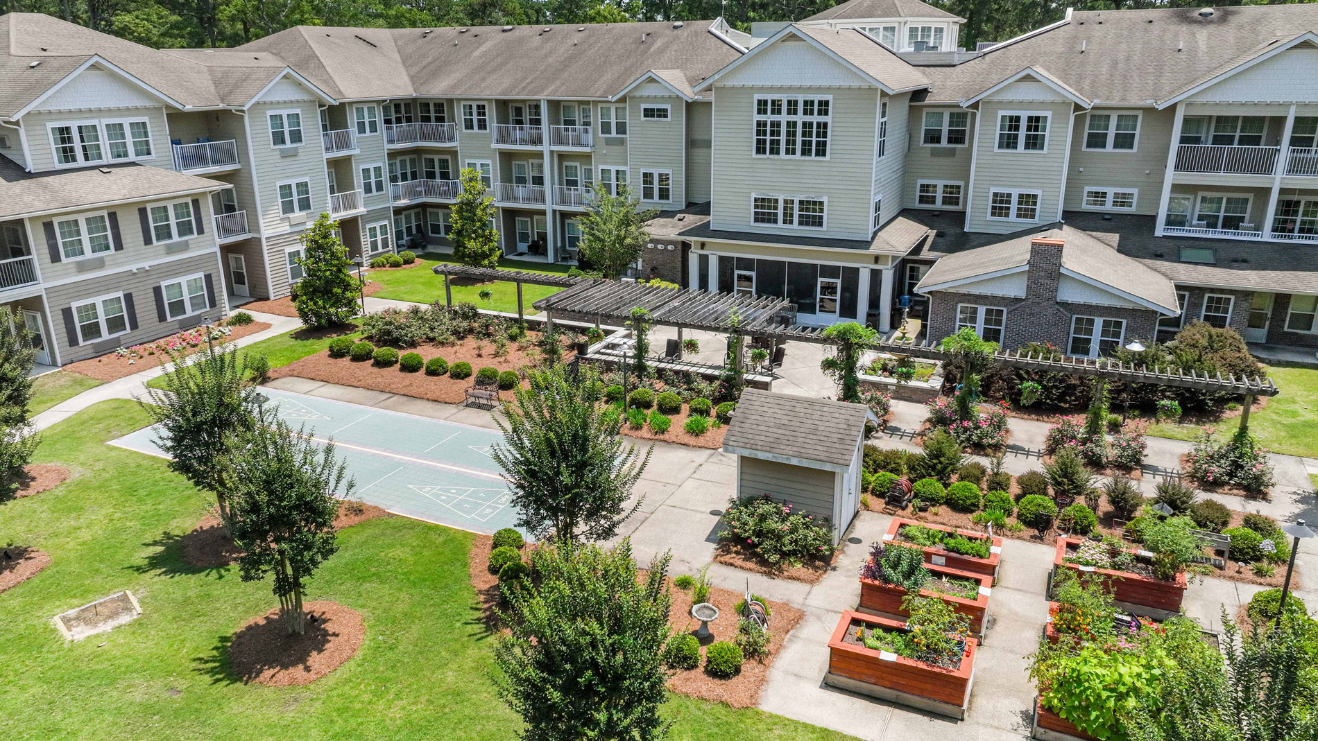 Aerial view of a modern apartment complex with three-story buildings, a landscaped courtyard, raised garden beds, a pergola, and a small sports court surrounded by trees and greenery.