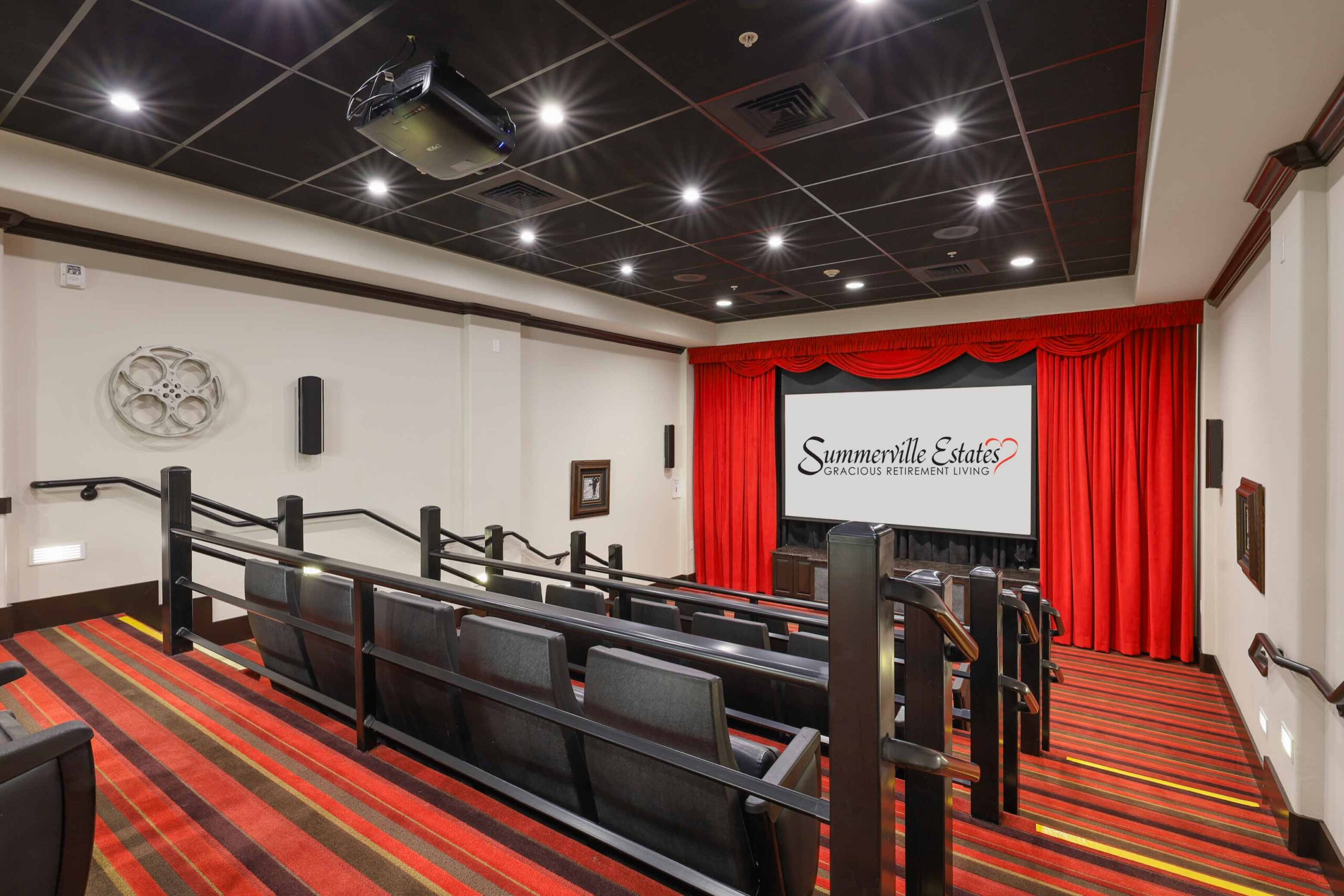 A small movie theater with tiered seating, red and black carpet, a projector on the ceiling, and a screen displaying "Summerville Estates Gracious Retirement Living" framed by red curtains.