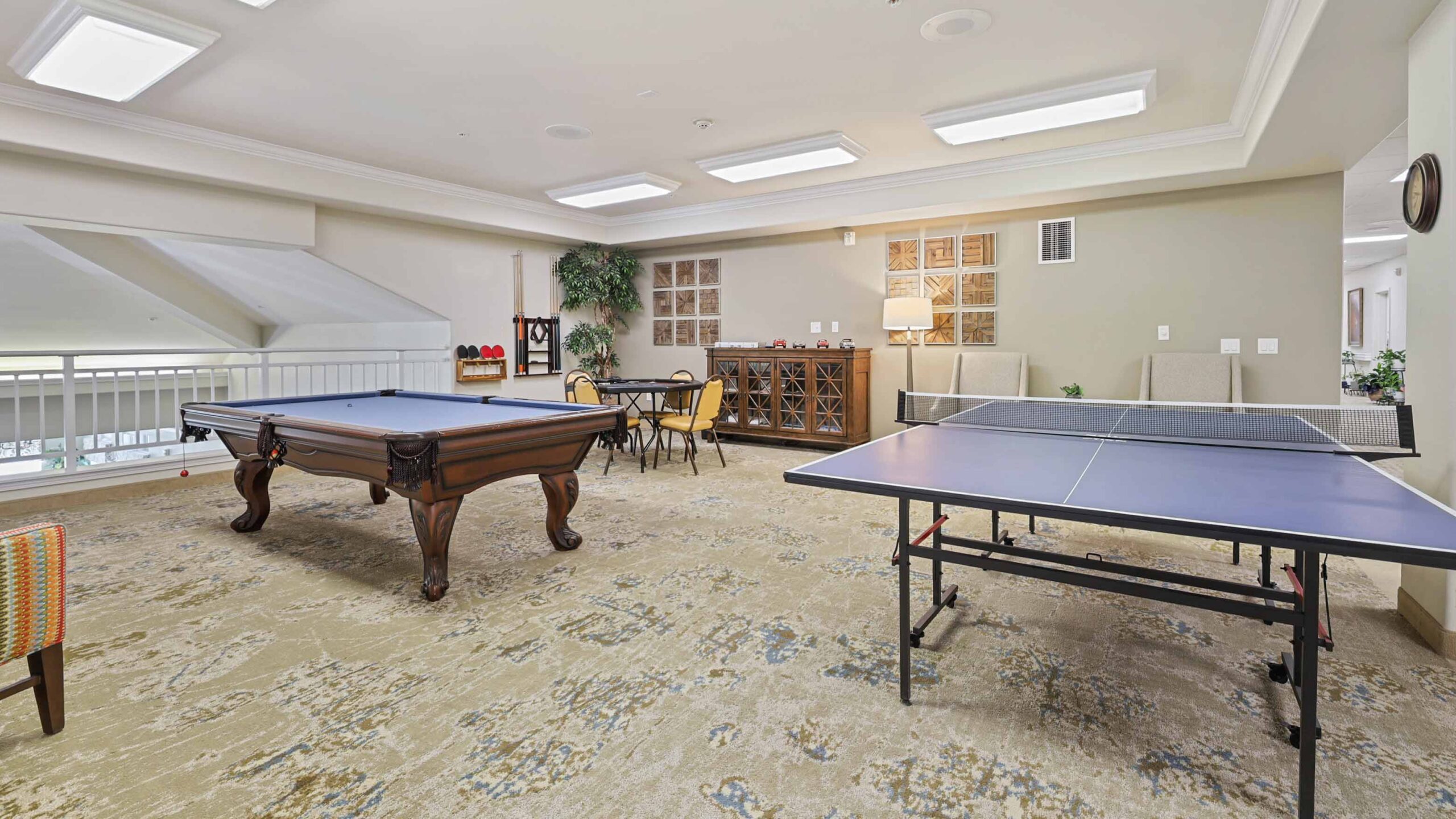 A spacious game room with a pool table and a ping pong table, cushioned chairs, a small round table with chairs, a wine rack, decorative wall art, and bright overhead lighting.