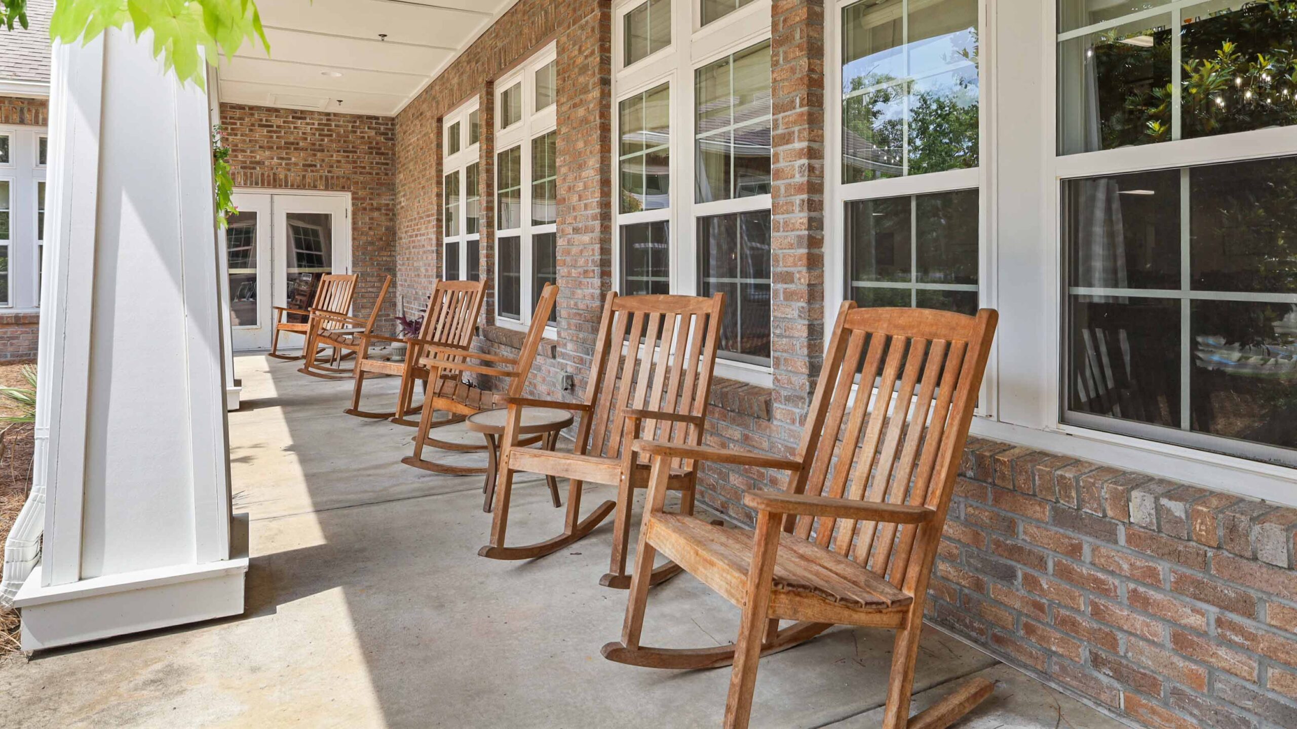 A row of wooden rocking chairs sits on a covered porch with brick walls and large windows, creating a peaceful and inviting outdoor space.