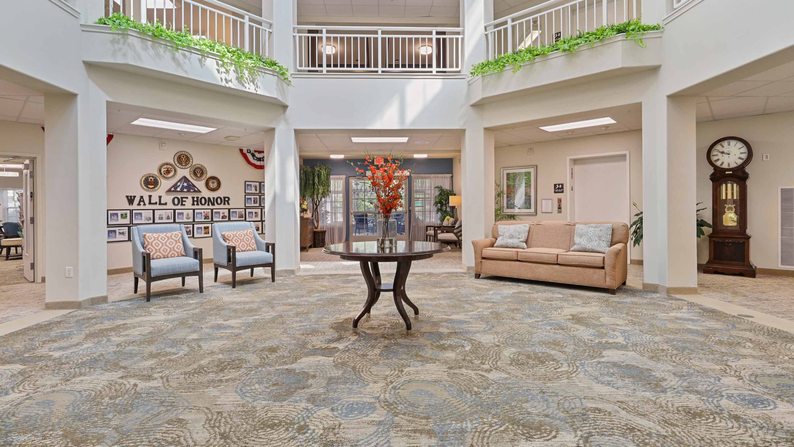 A bright, spacious lobby with a round table and orange floral arrangement at the center, surrounded by armchairs, a sofa, a wall clock, potted plants, and a “Wall of Honor” display. White railing and natural light above.