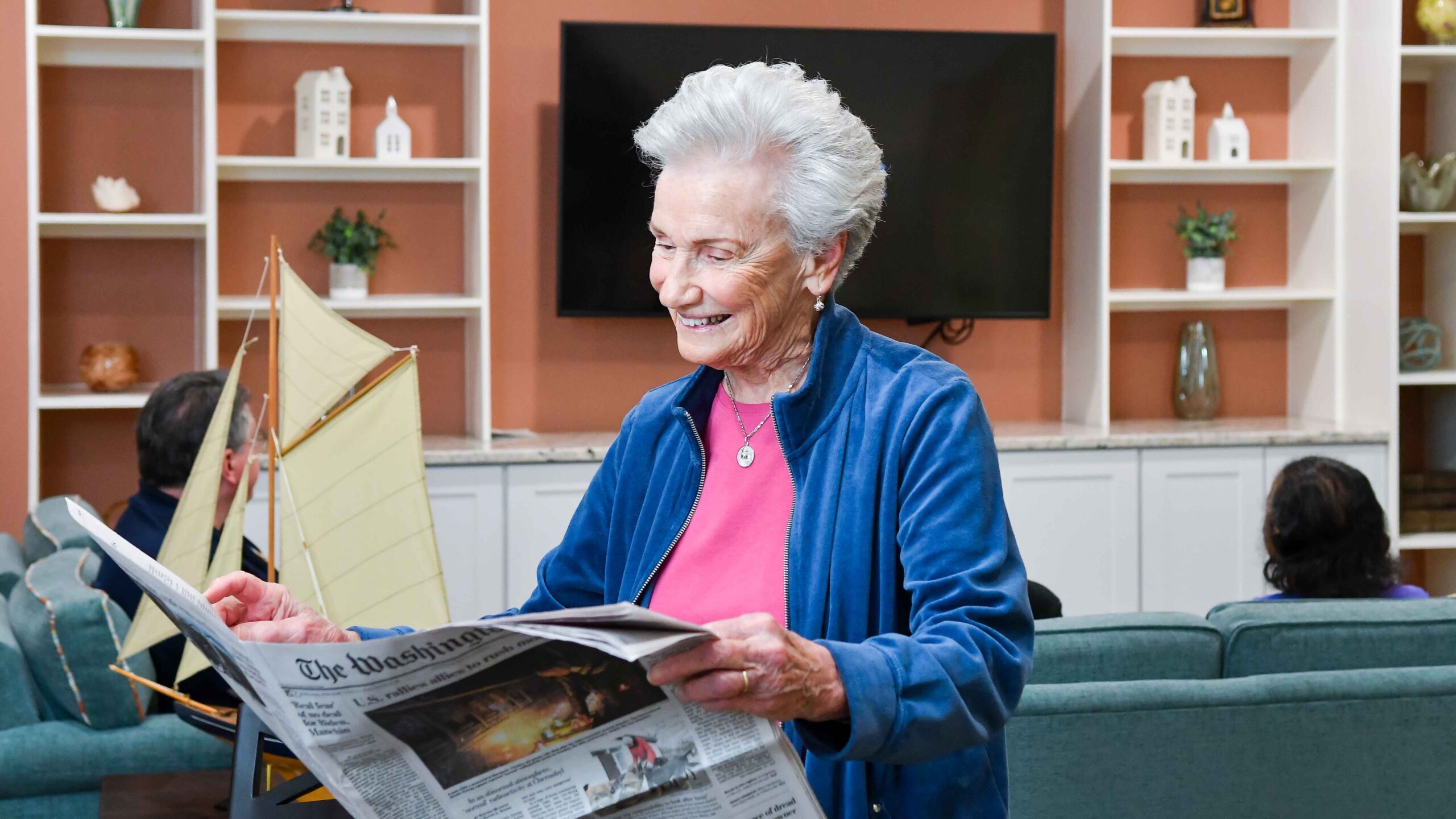 An elderly woman with short gray hair smiles while reading a newspaper in a cozy living room with shelves and a model sailboat in the background. Other people are seated on couches behind her.