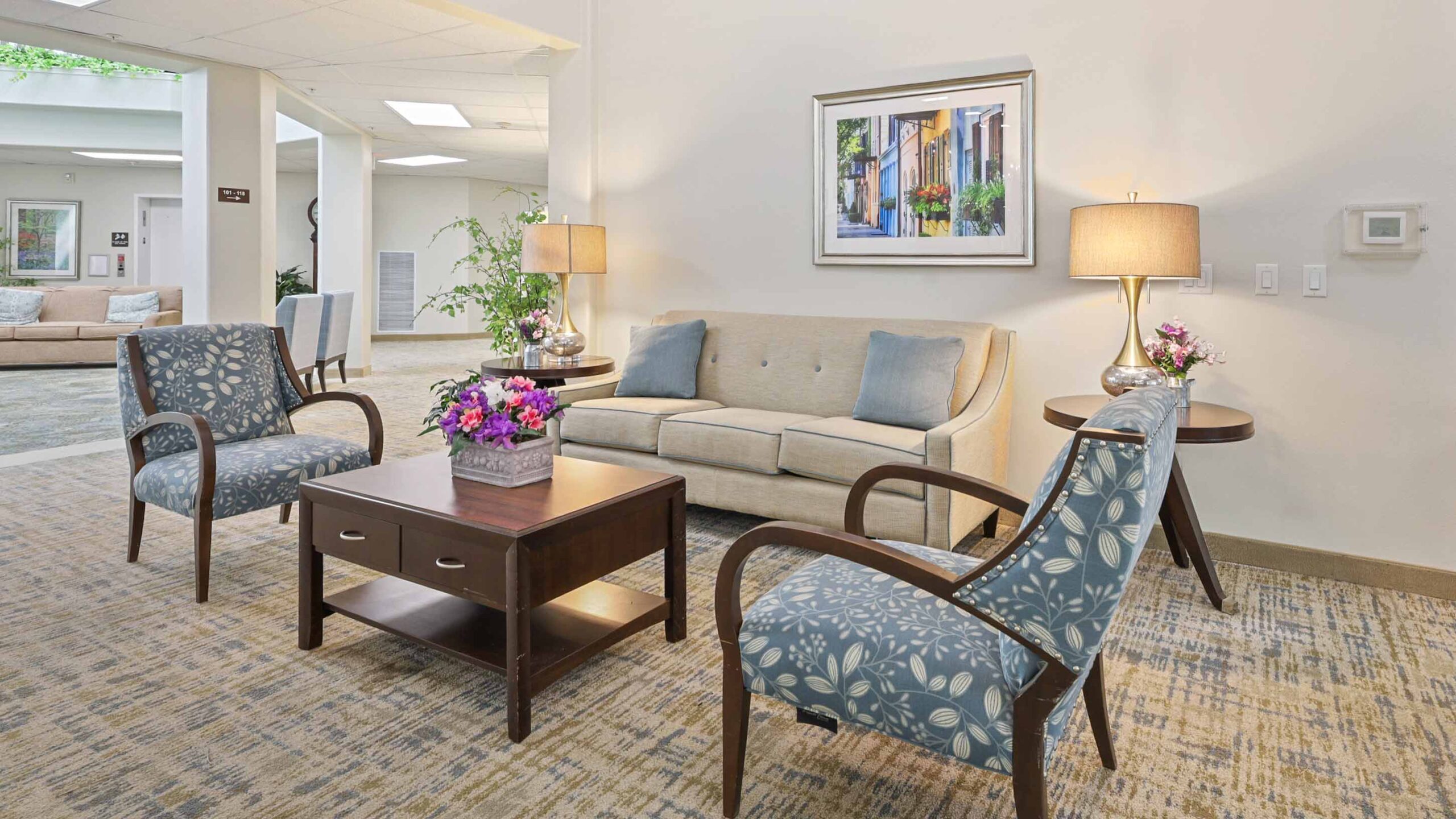 A cozy lounge area with a beige sofa, two blue patterned armchairs, a wooden coffee table with flowers, two side tables with lamps, a wall painting, and light-colored walls and carpet.