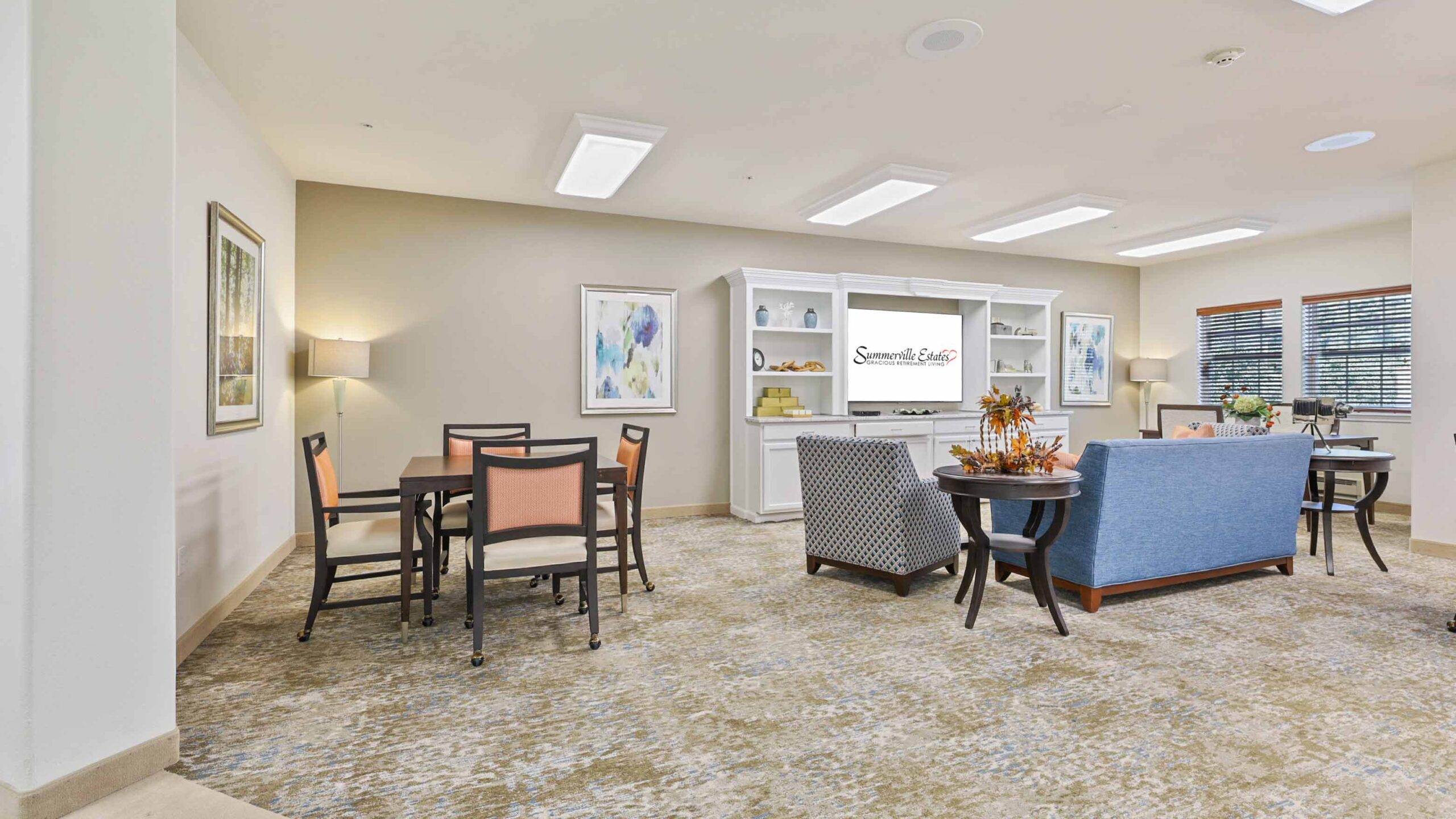 A bright, spacious room with a dining table and chairs, a blue couch, armchairs, artwork on the walls, a bookshelf, and decorative plants, creating a cozy, inviting atmosphere.