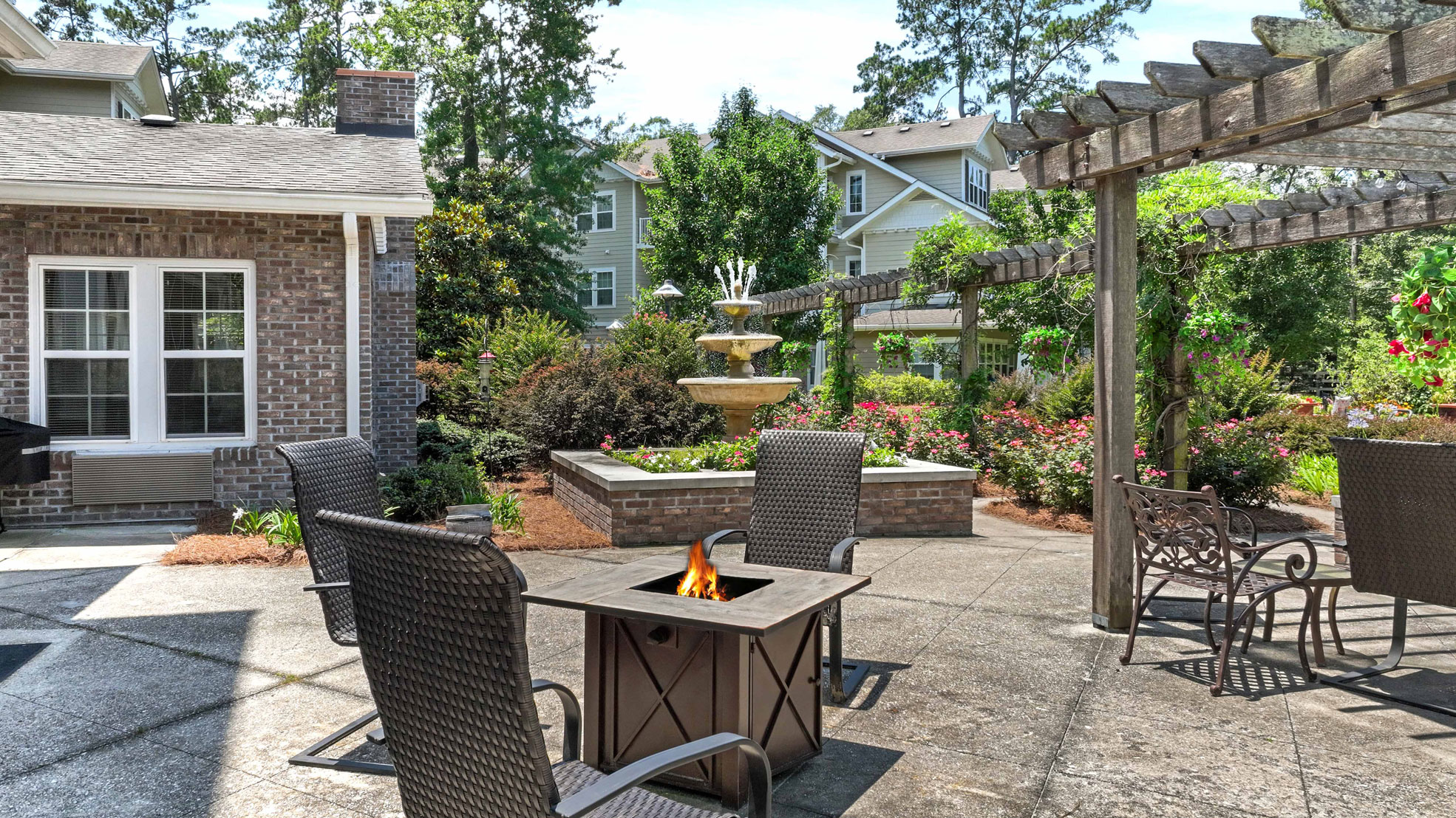 A sunny backyard patio with wicker chairs around a small fire pit table, a brick house, lush greenery, blooming flowers, a pergola, and a tiered water fountain in the background.