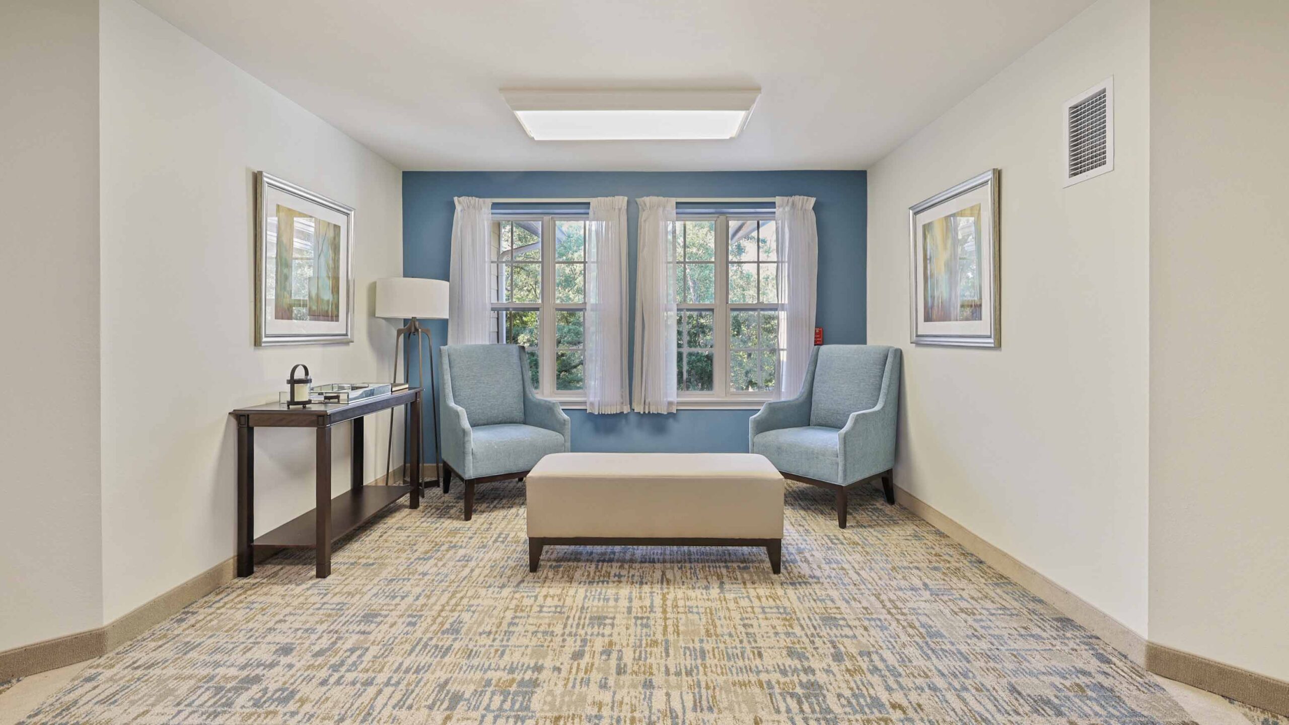 A small, bright seating area with two blue armchairs, a bench, a side table with a lamp and tray, two windows with white curtains, framed art on the walls, and patterned carpet.