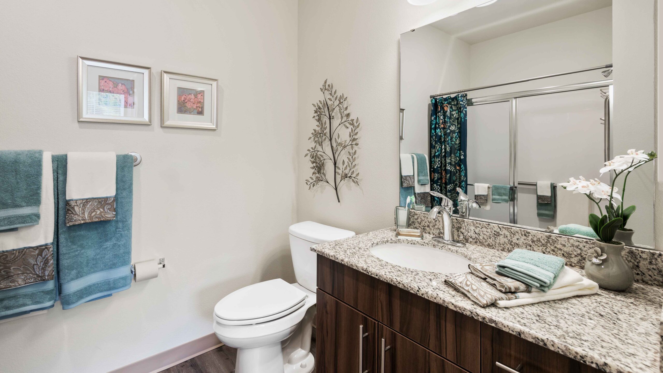 Modern bathroom with a granite countertop, dark wood vanity, towels on a rack, toilet, wall art, framed pictures, a potted orchid, and a shower with glass doors and a blue floral curtain.