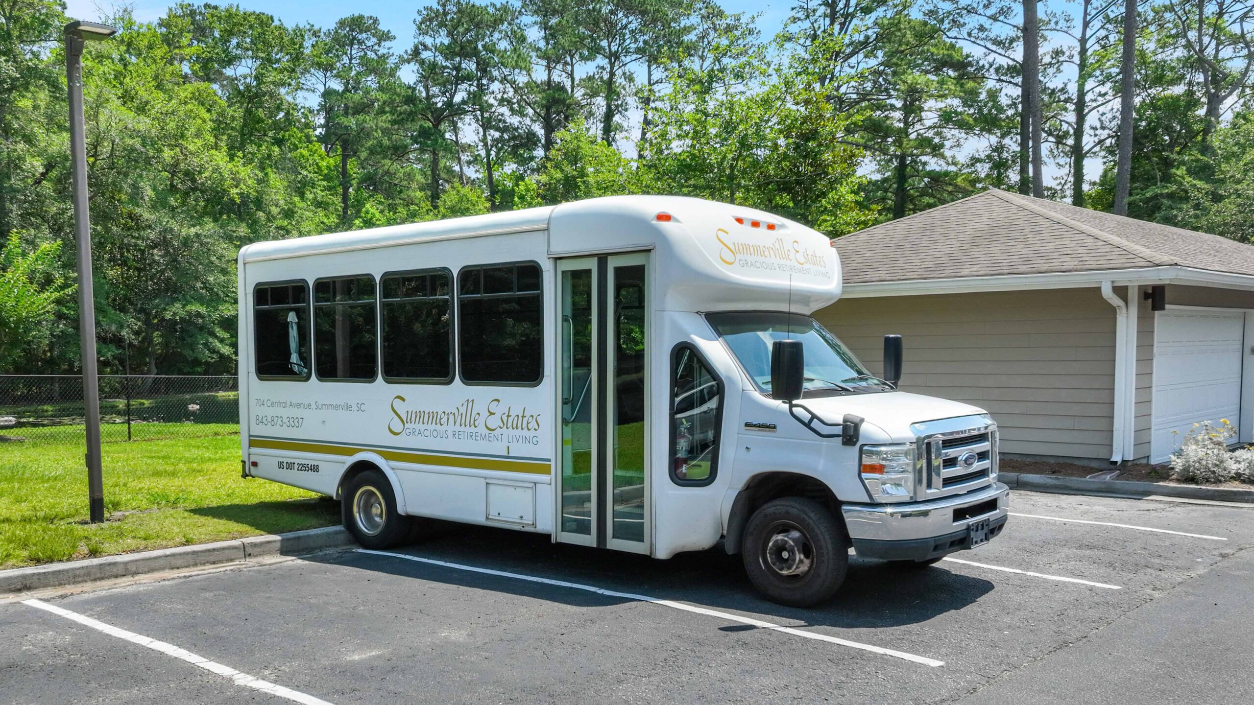 A white shuttle bus labeled “Summerville Estates” is parked in an asphalt lot beside a tan building, with green trees and grass visible in the background on a sunny day.