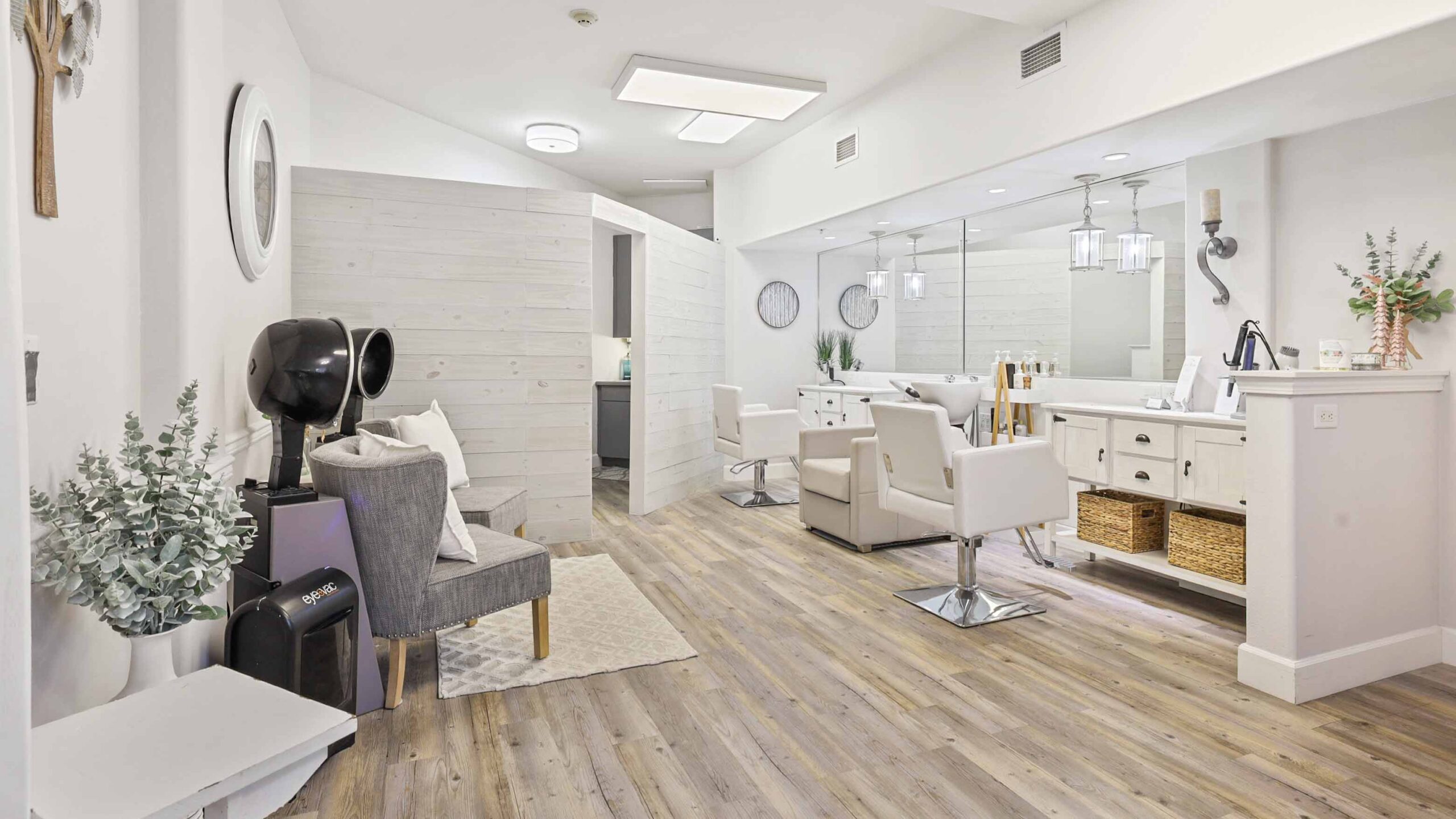 A modern, bright hair salon with white walls and wooden floors. There are white styling chairs, large mirrors, and a cozy waiting area with a gray armchair and potted plants. The space appears clean and inviting.