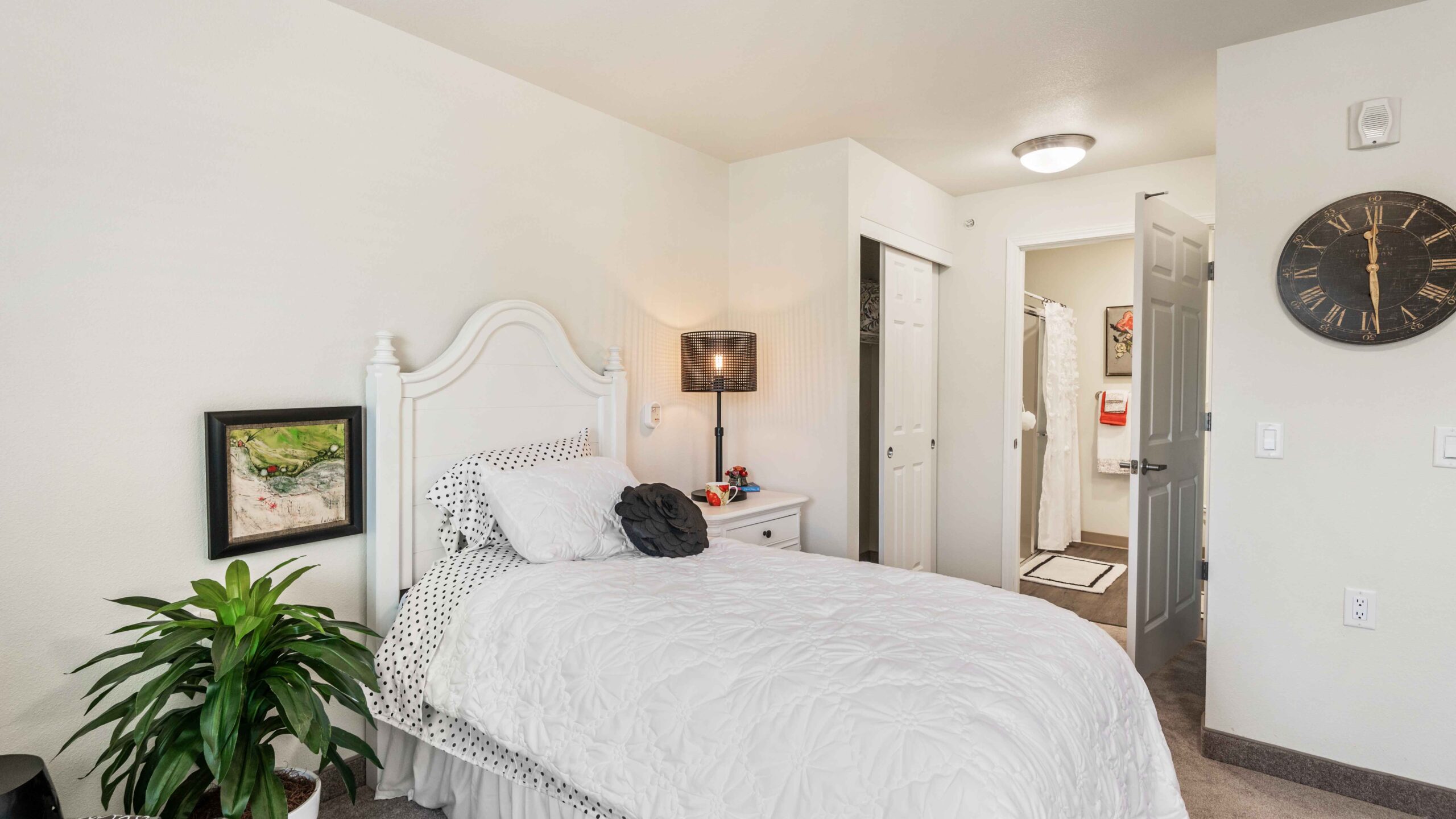 A cozy bedroom with a single white bed, polka-dot bedding, a nightstand with a lamp, a potted plant, and a large black wall clock. An open door reveals a bathroom with white towels and decor.