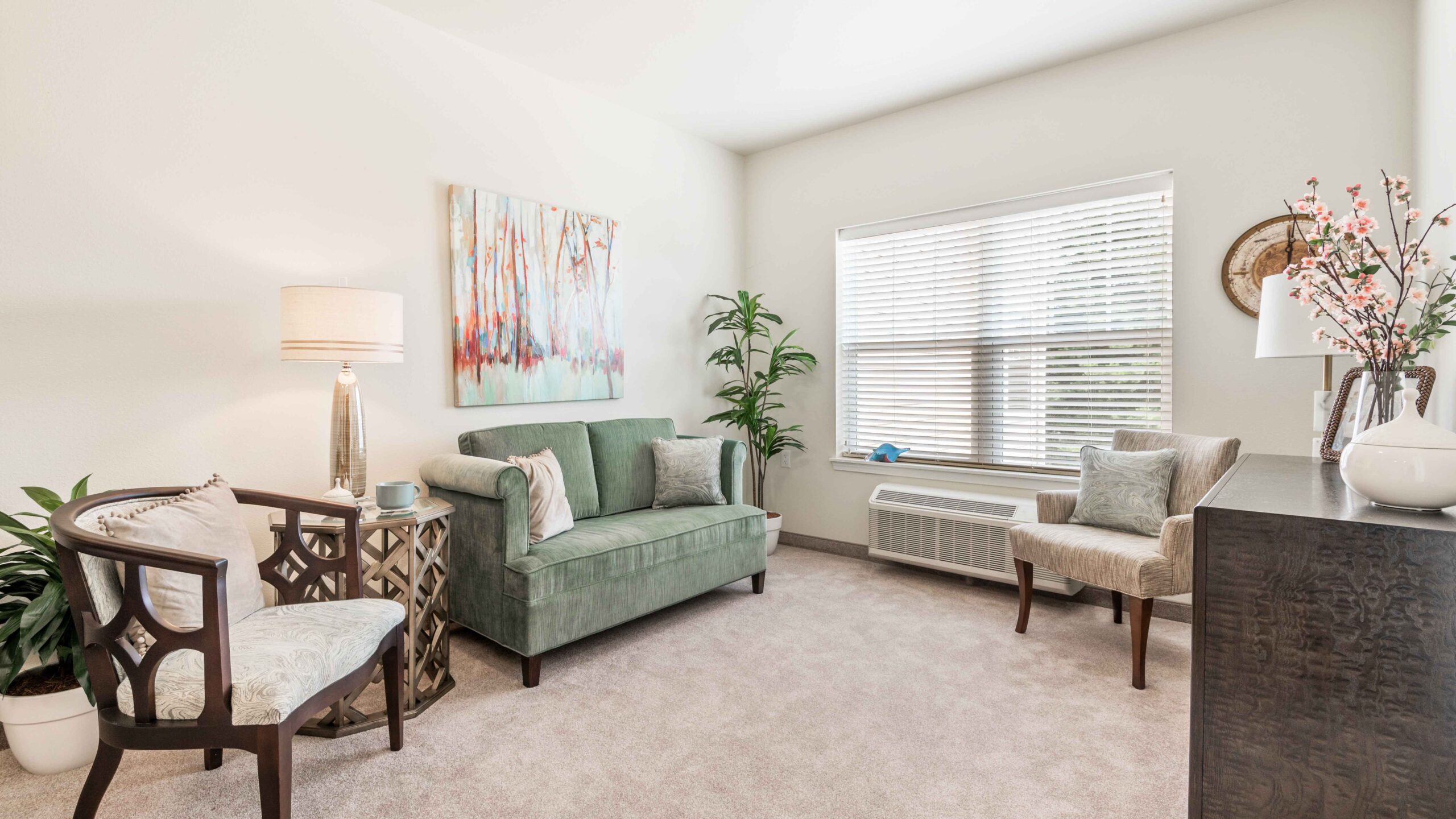 Bright, cozy living room with a green loveseat, two armchairs, a lamp, side table, large window with blinds, plants, and soft, neutral decor. A colorful abstract painting hangs above the loveseat.