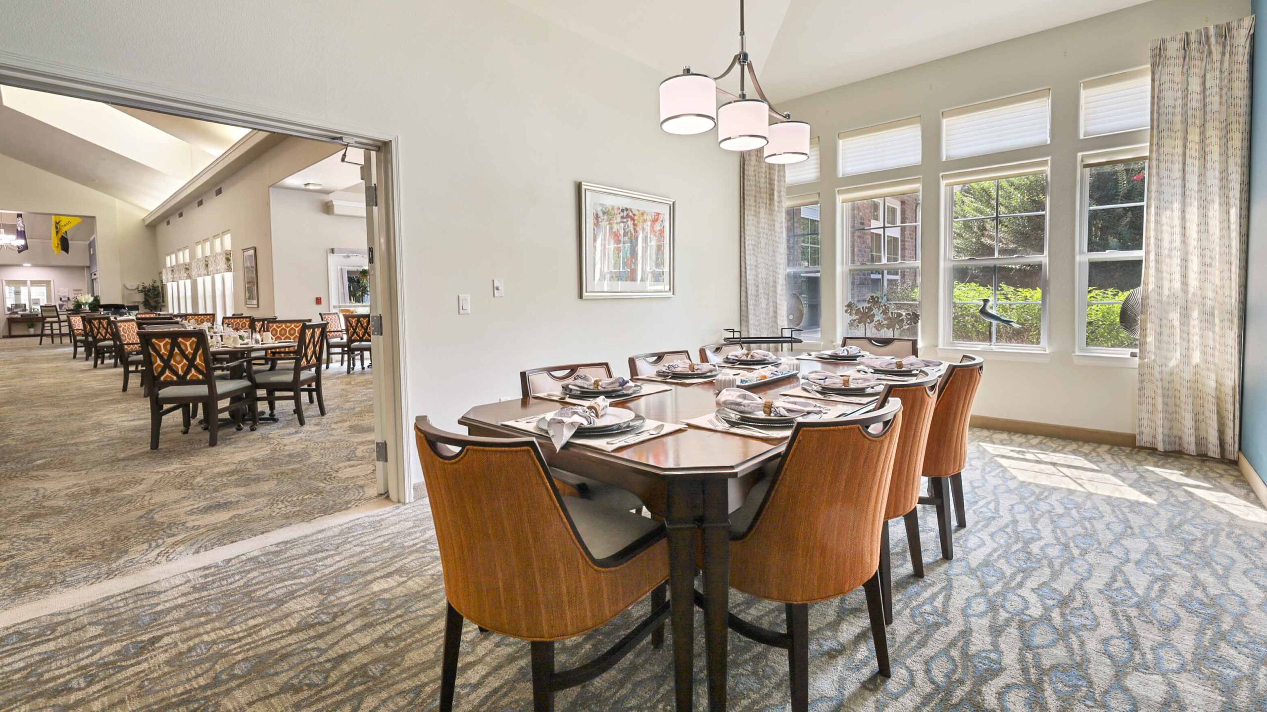 A bright dining area with a set table for eight, wooden chairs, large windows letting in natural light, modern light fixtures, and an adjacent larger dining room visible through an open doorway.