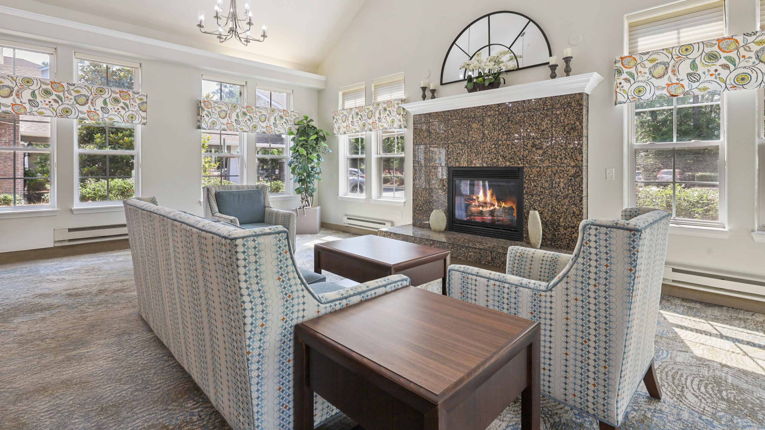 Bright living room with patterned armchairs and a sofa arranged around a coffee table near a lit fireplace, large windows with colorful valances, a chandelier, and decorative plant in the corner.