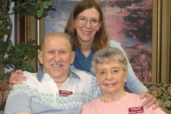 Three smiling individuals, two elderly adults seated on a floral-patterned couch and a middle-aged woman standing behind them, all wearing name tags. They are indoors next to green plants and a wall featuring abstract artwork in warm colors.