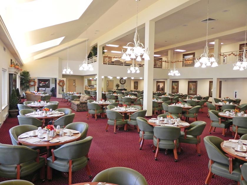 A spacious dining area with high ceilings, featuring several round tables set for meals with green upholstered chairs. The room is well-lit with modern chandeliers and has a red carpet. In the background, there are couches, a staircase, and various wall decorations.