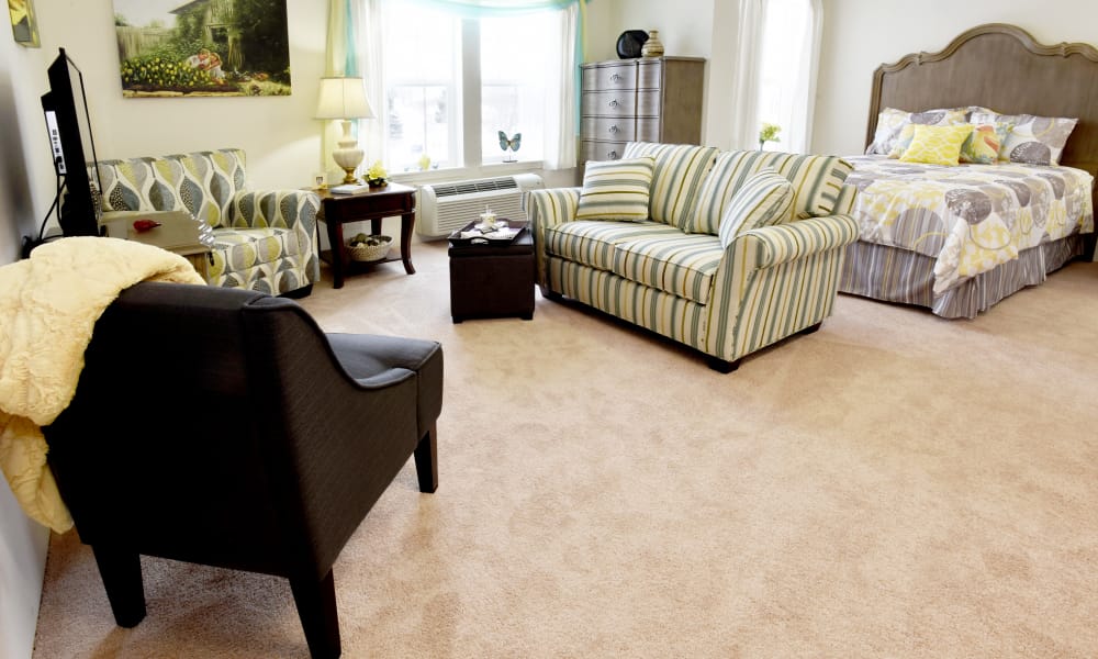 A well-lit room with beige carpet features a bed with floral bedding, a striped loveseat and chair, a black armchair, a small table with a lamp, and a wall-mounted TV. A dresser and a window with green curtains are also visible. Decor includes a framed painting.