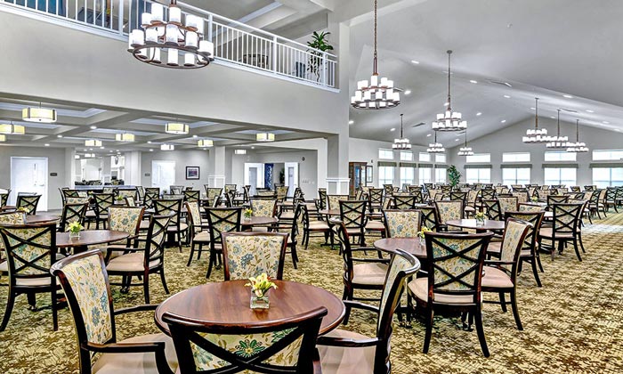 A spacious, well-lit dining area with numerous round tables and cushioned chairs neatly arranged. Chandeliers hang from the high ceiling, and large windows allow natural light to flood the room, giving it a bright and airy atmosphere.