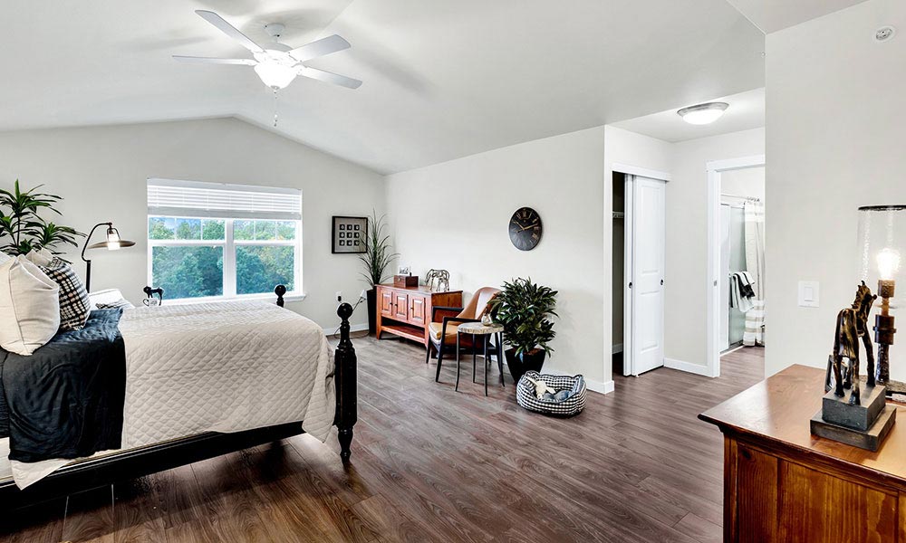 A spacious, modern bedroom with a vaulted ceiling and large window. The room features a bed with white and black bedding, a wooden floor, a ceiling fan, a small study area with a desk, and a plant. The attached bathroom is visible through an open doorway.