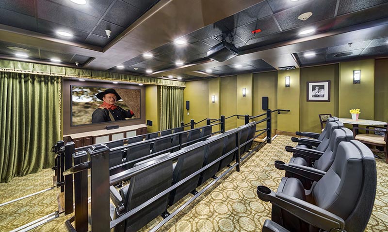 A cozy home theater room featuring tiered seating with black leather chairs, a large screen showing a person in a cowboy hat, green curtains framing the screen, and a patterned carpet. Wall sconces provide ambient lighting, and a framed picture hangs on the wall.