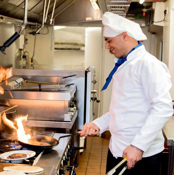 A chef in a white uniform and hat is cooking in a professional kitchen. He is smiling while flambéing in a skillet on a stove, producing visible flames. The kitchen has stainless steel appliances and appears clean and organized.