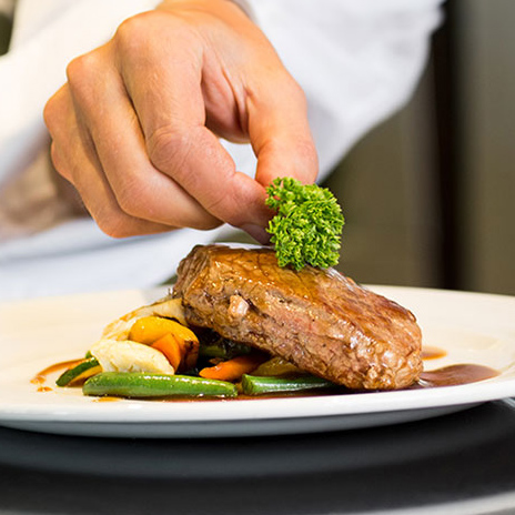 A chef's hand is garnishing a cooked steak with a sprig of fresh parsley. The steak is plated on top of sautéed vegetables, including green beans and yellow bell peppers, drizzled with a savory brown sauce, all presented on a white plate.