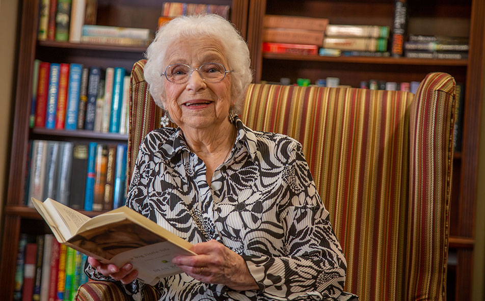 An elderly woman with white hair and glasses sits on a striped armchair, holding an open book and smiling. Behind her, a wooden bookshelf filled with colorful books is visible. She is wearing a patterned blouse.