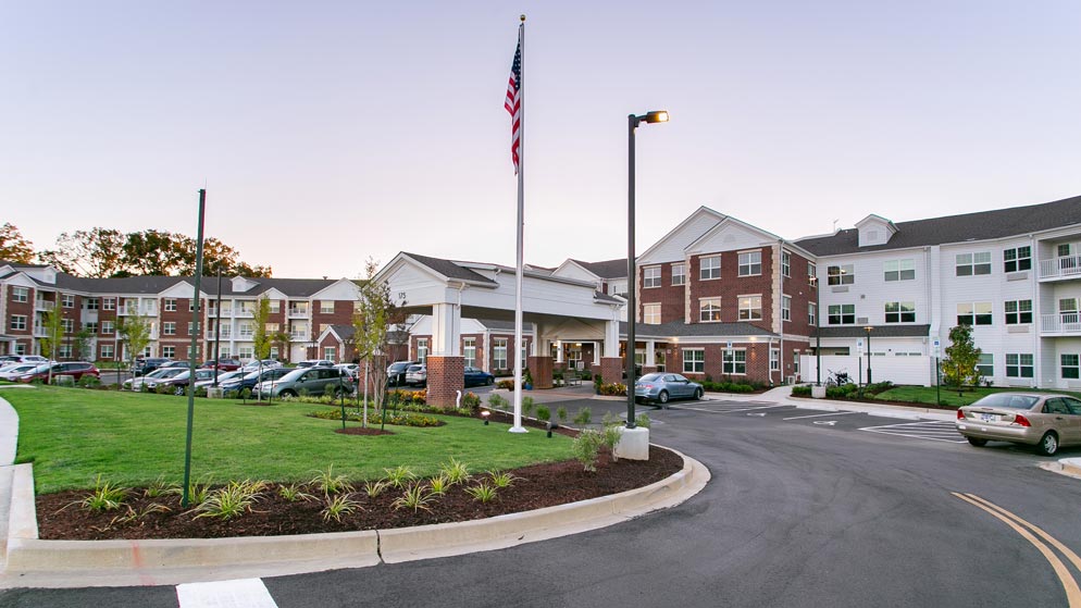 A large residential building complex with a mix of brick and white siding. There is a paved circular driveway, an American flag on a flagpole, several parked cars, and well-maintained green lawns with small shrubs.