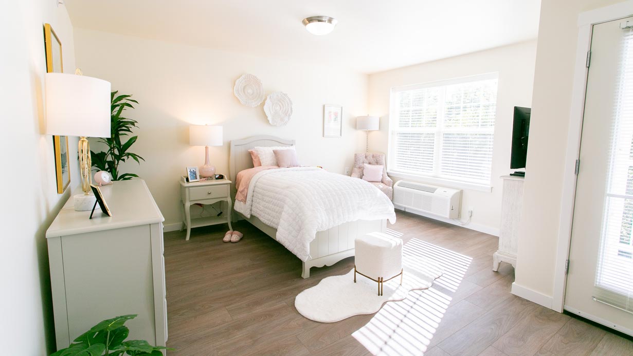 Bright, airy bedroom with a neatly made bed in white bedding, two side tables with lamps, a dresser with a plant, and a soft rug. Sunlight streams through large windows, highlighting the minimalist decor and cozy atmosphere.