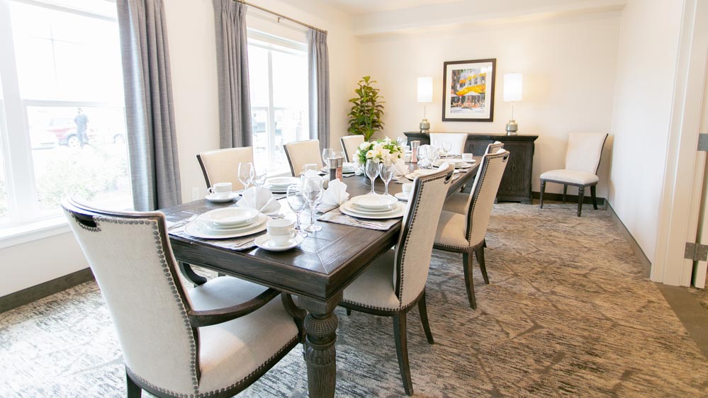 A well-lit dining room with a long, dark wood table set for eight, featuring white plates and glassware. Light upholstered chairs surround the table. A large window with gray curtains brightens the space, along with two lamps on a sideboard.