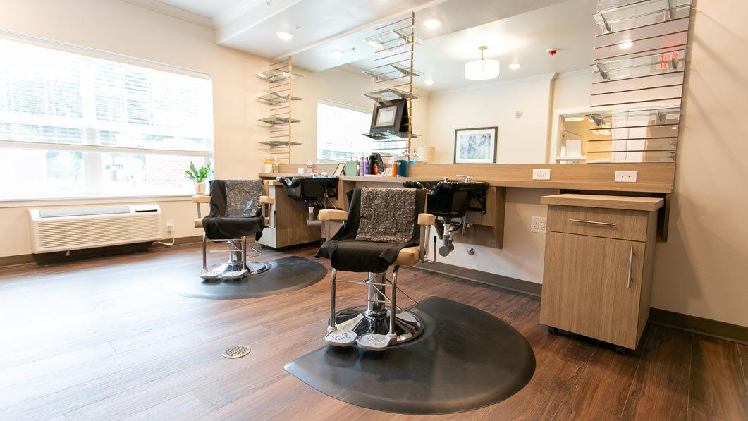 A modern hair salon with two styling chairs and mirrors. The space has wooden flooring, shelves with styling products, and bright natural light from large windows. The decor is clean and minimalistic.