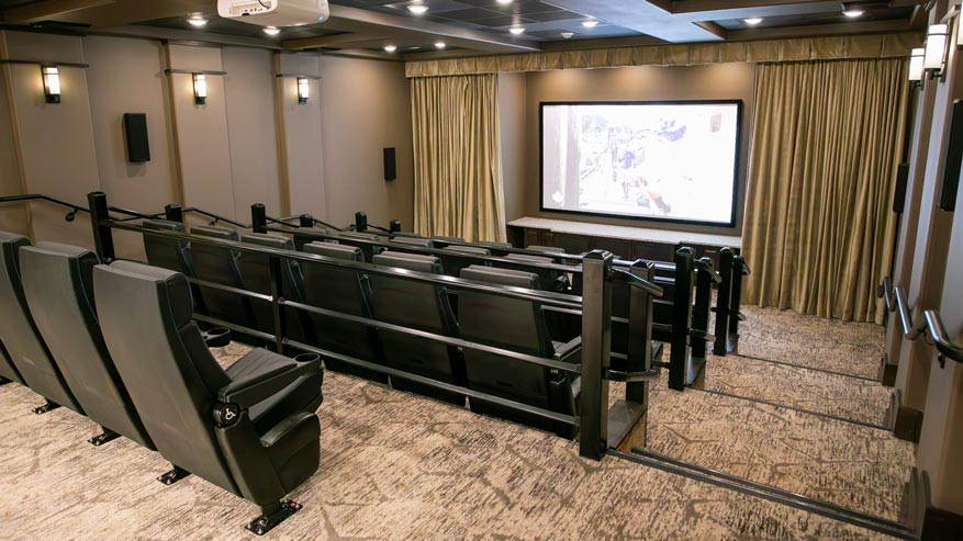 A home theater with tiered seating, equipped with black chairs facing a large screen. The room features beige walls, soft lighting, and heavy curtains flanking the screen.