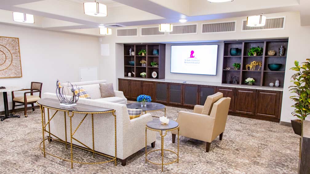 A cozy living room with a light gray sofa and two beige armchairs arranged around a circular coffee table. A large TV is mounted on a brown shelving unit displaying the St. Jude Children's Research Hospital logo. Decorative plants and art adorn the space.