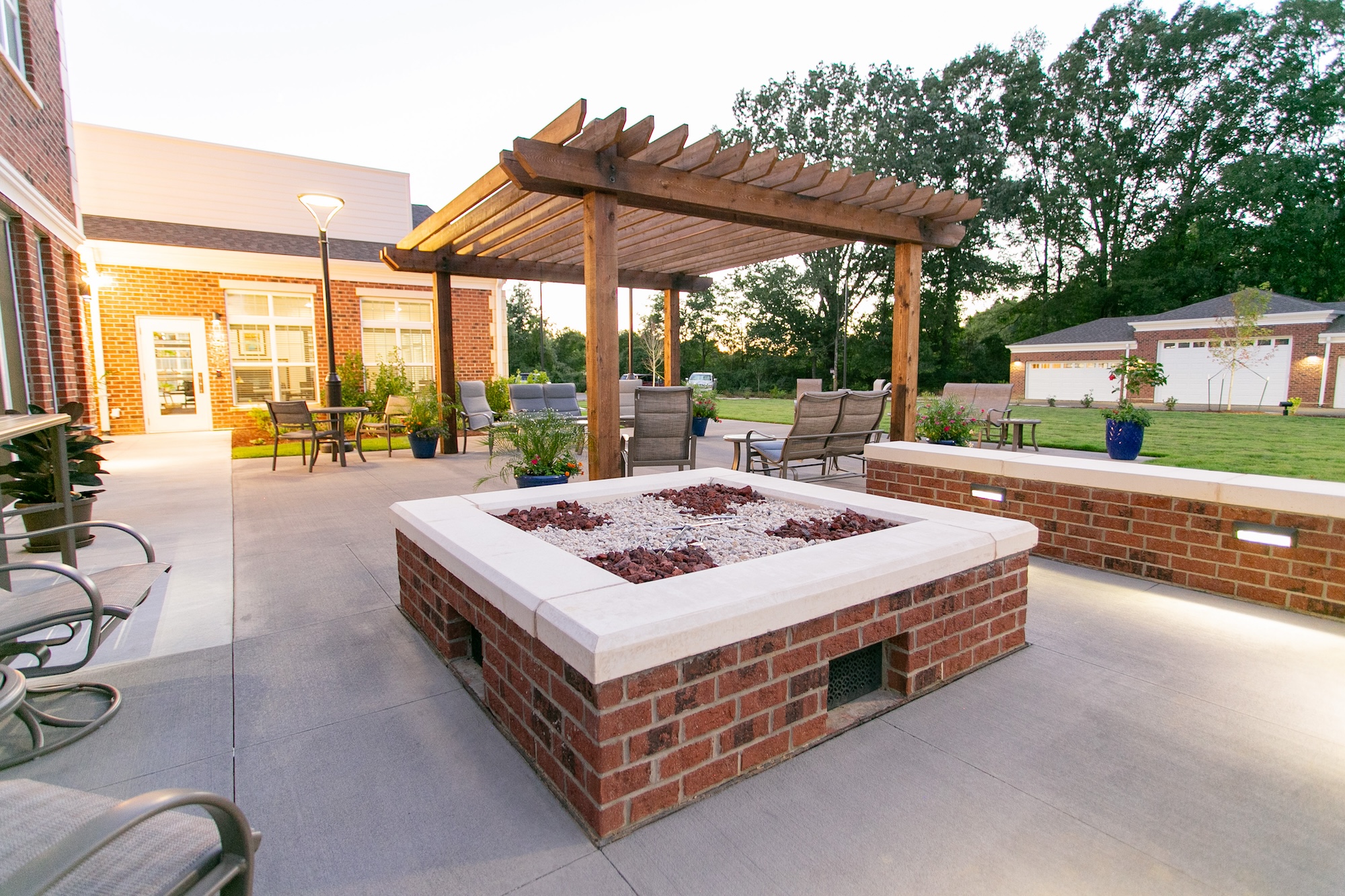 Outdoor patio with a large brick fire pit in the center, surrounded by patio chairs. A wooden pergola provides partial shade. The area is bordered by lush trees, and a grassy lawn is visible in the background.