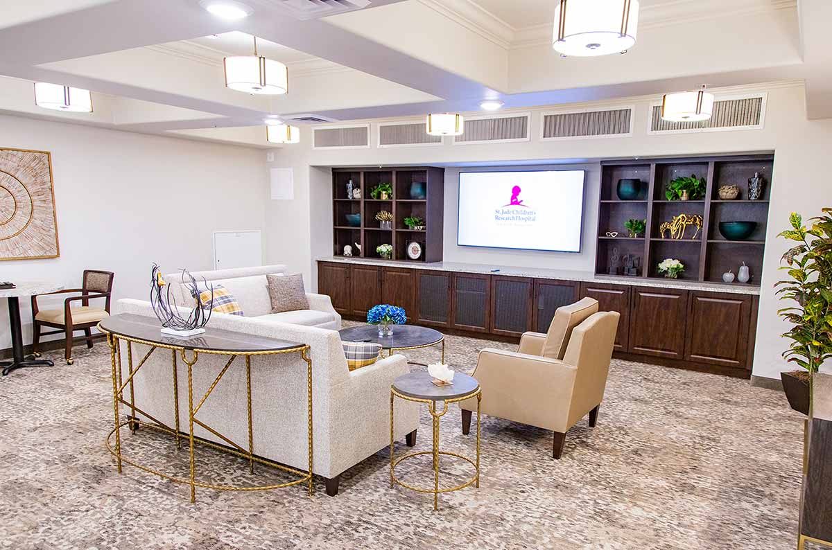 A stylish waiting room features seating with beige sofas and tan armchairs around a coffee table. A large TV is mounted on built-in shelves displaying a logo. The room is warmly lit, with decorative plants and artwork enhancing the cozy atmosphere.