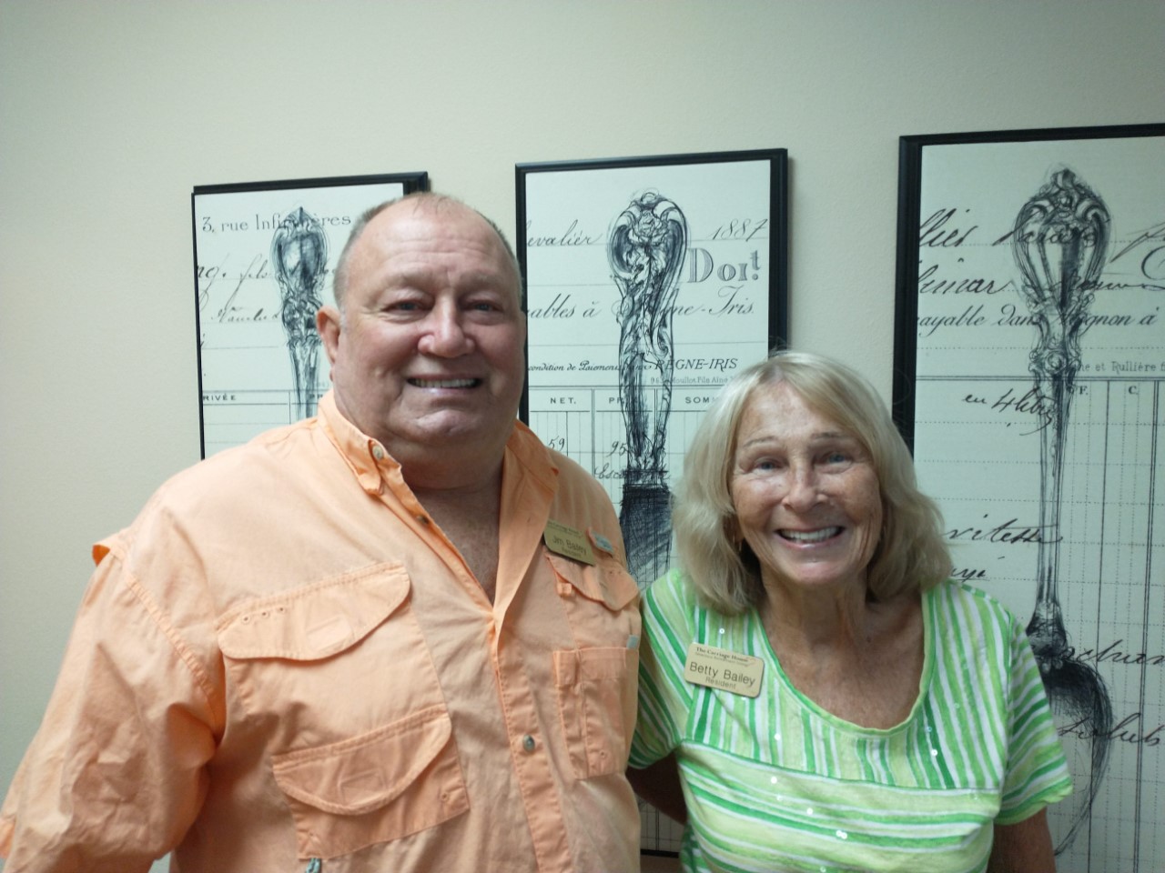 A man in an orange shirt and a woman in a green striped shirt stand smiling in front of framed artwork on a wall. Both are wearing name tags. The man is on the left, and the woman is on the right.