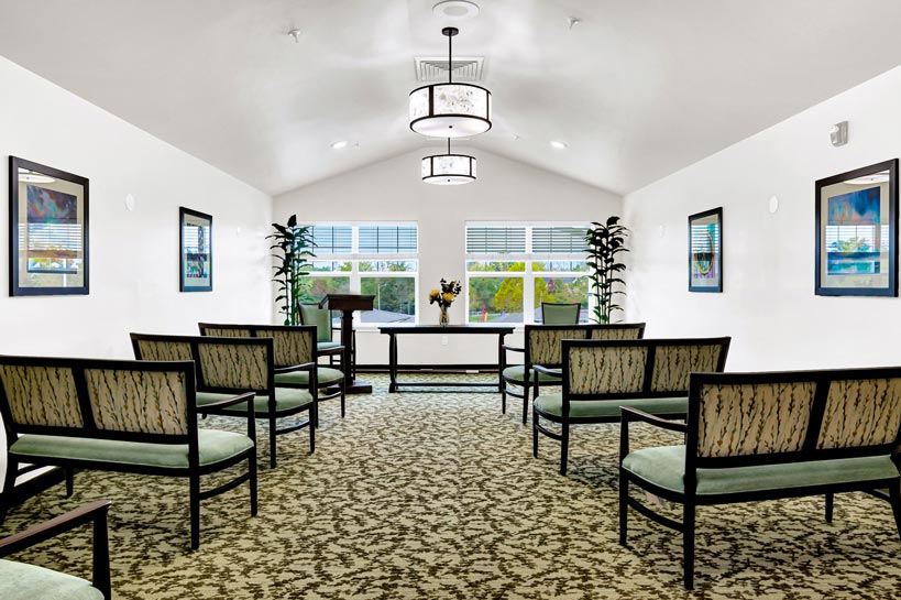 A small, well-lit meeting room with a high ceiling features rows of padded chairs facing a lectern. Large windows along the back wall let in natural light, while plants and framed artwork adorn the white walls. The patterned carpet adds a touch of elegance.