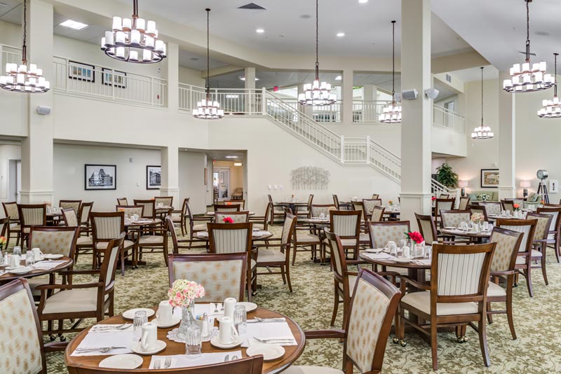 An elegant dining area with neatly arranged tables and chairs. The tables are set with white plates, cups, and cutlery, and decorated with small flower arrangements. There is a staircase leading to a mezzanine level, and multiple chandeliers hanging from the ceiling.